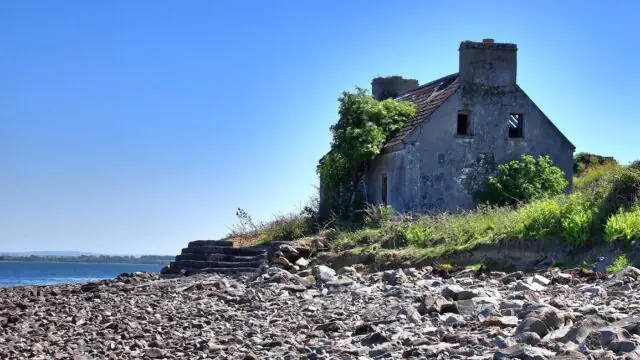 An old, abandoned stone house with a damaged roof sits by a rocky shoreline under a clear blue sky.
