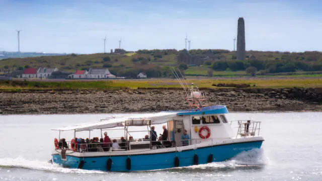 A small passenger ferry with people onboard sails near a rocky shoreline, with houses, wind turbines, and a tall stone tower visible in the background.