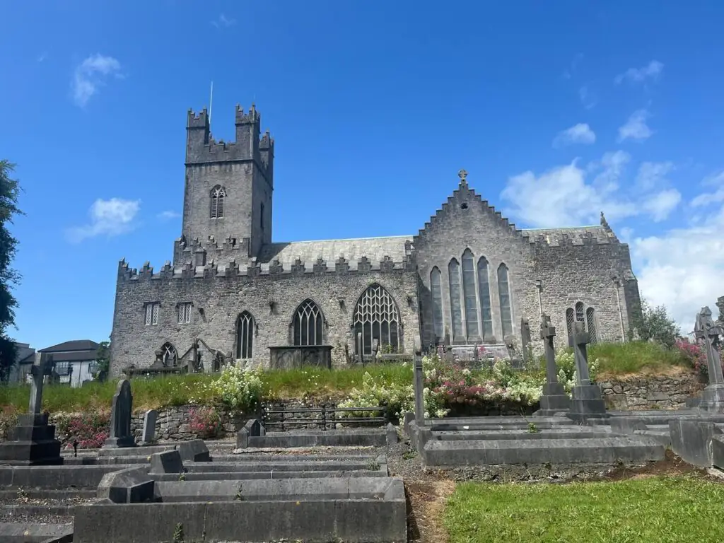 Stone church with a tall square tower, arched windows, and gravestones in the foreground under a partly cloudy blue sky.
