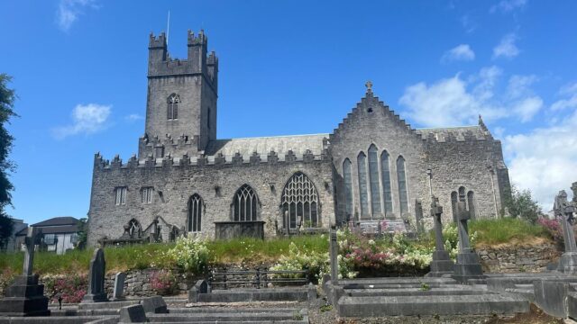 Stone church with a tall square tower, arched windows, and gravestones in the foreground under a partly cloudy blue sky.