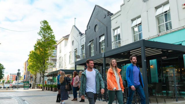 Three people walk in Limerick city street lined with modern buildings, while other pedestrians and outdoor diners.