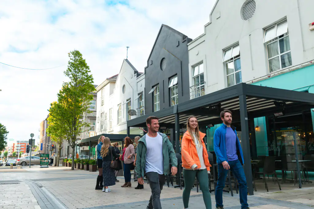 Three people walk on a city street past outdoor seating and other pedestrians, with modern buildings and trees lining the sidewalk under a partly cloudy sky.