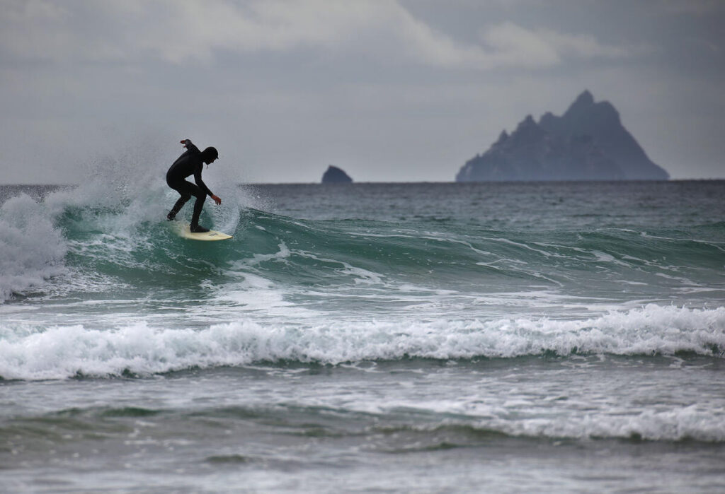A surfer in a wetsuit rides a small wave near the shore, with rocky islands visible in the distance under a cloudy sky.