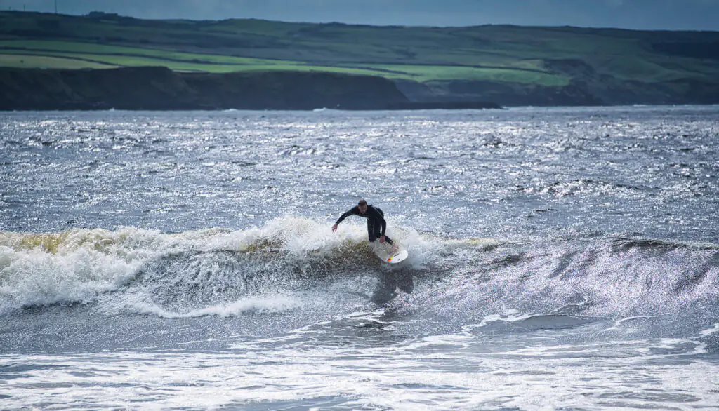 A surfer in a wetsuit rides a wave near the shoreline with green cliffs in the background under a cloudy sky.