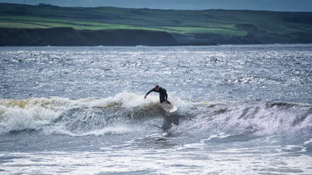 A surfer in a wetsuit rides a wave near the shoreline with green cliffs in the background under a cloudy sky.