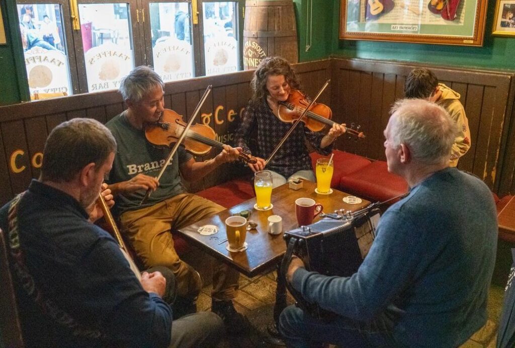 Five people sit around a table in a pub, playing traditional musical instruments including fiddles and an accordion, with drinks on the table.