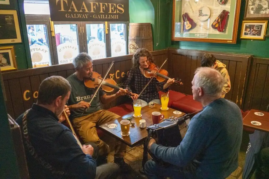 Five people play traditional Irish music on fiddles and an accordion at a wooden table in a cozy pub with drinks on the table.