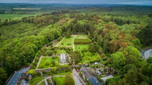 Aerial view of a formal garden with geometric hedges, lawns, and several buildings, surrounded by dense forest and countryside.