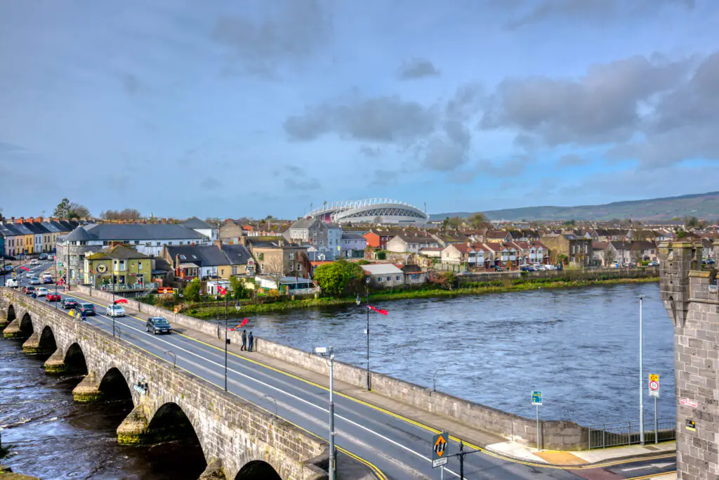 A stone bridge spans a river beside a row of houses, with a large stadium visible in the background under a partly cloudy sky.