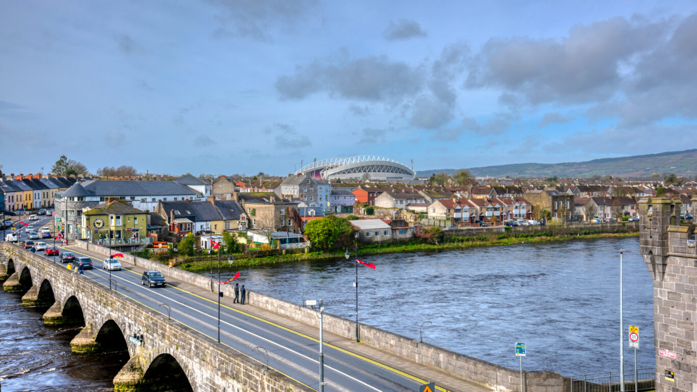 A stone bridge spans a river beside a row of houses, with a large stadium visible in the background under a partly cloudy sky.
