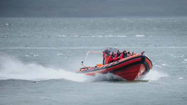 A red inflatable speedboat with several people in life jackets moves quickly across the water, creating a spray behind it.
