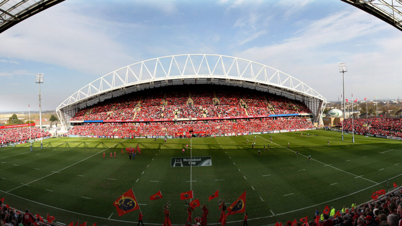 Wide-angle view of a packed rugby stadium with fans in red, a game in progress on the field, and clear skies overhead.