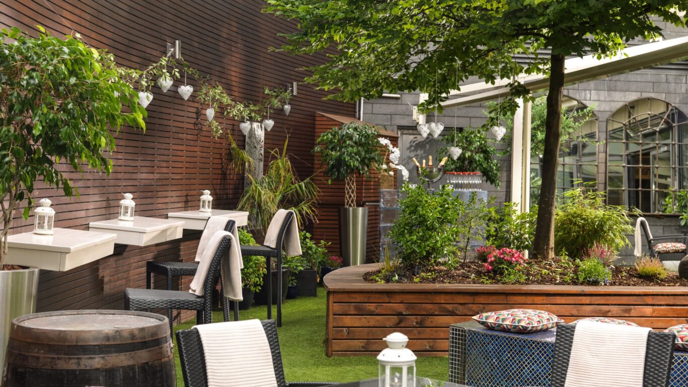 Outdoor patio with wicker chairs, glass tables, white lanterns, potted plants, wooden panel walls, and a raised garden bed with a tree and flowers.