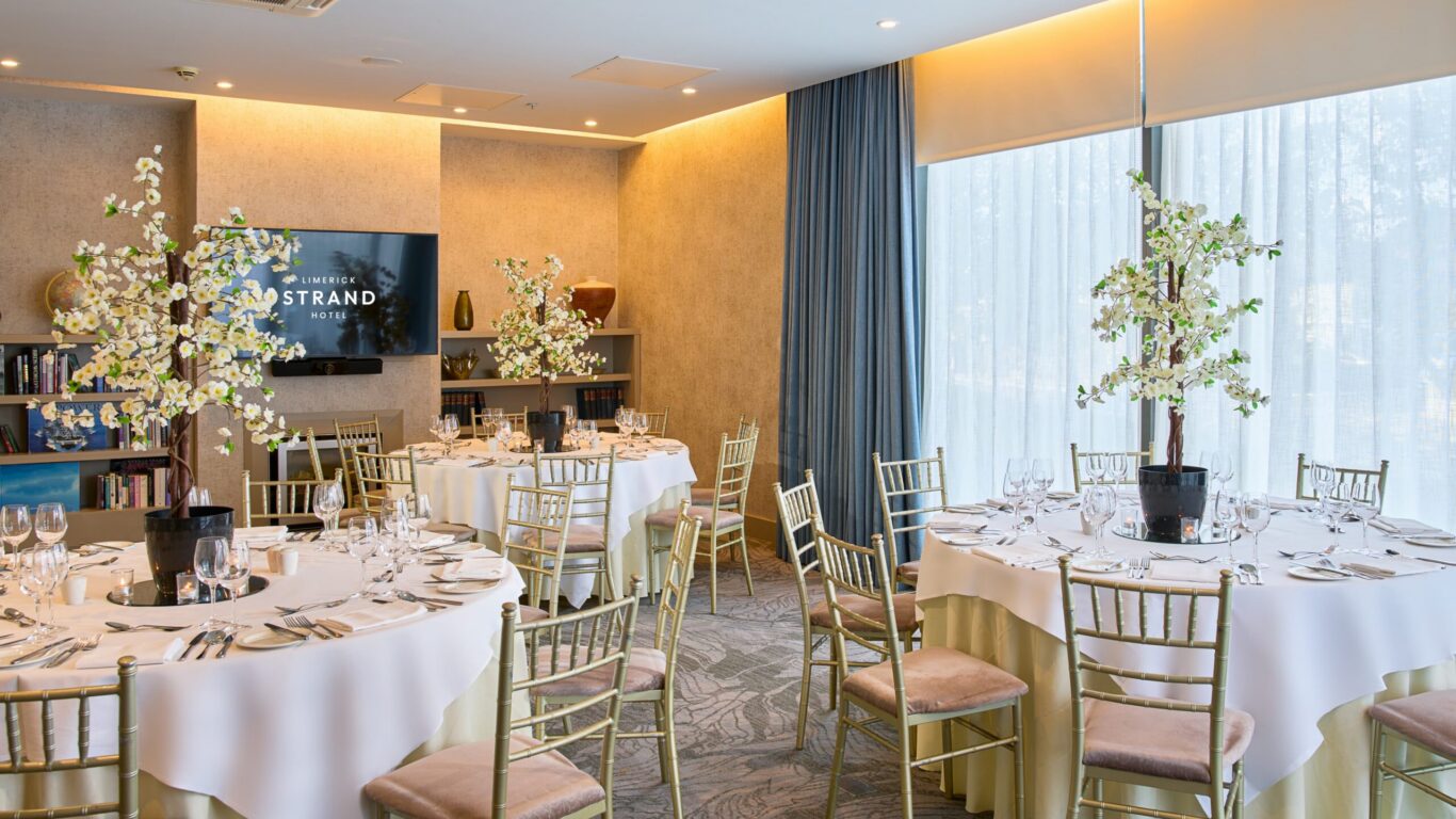 A banquet room with round tables set for dining, gold chairs, white tablecloths, floral centerpieces, and a TV screen displaying 