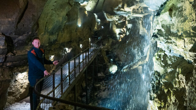 A person stands on a metal walkway inside a cave, next to a rock wall with water cascading down and illuminated by lights.