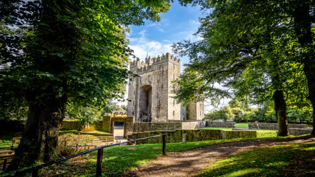 A stone castle with a central tower stands behind trees and a stone wall on a bright, sunny day with blue sky and clouds.