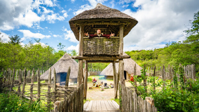 A person leans out from a raised wicker hut surrounded by a wooden fence, with round thatched-roof houses and greenery in the background under a partly cloudy sky.