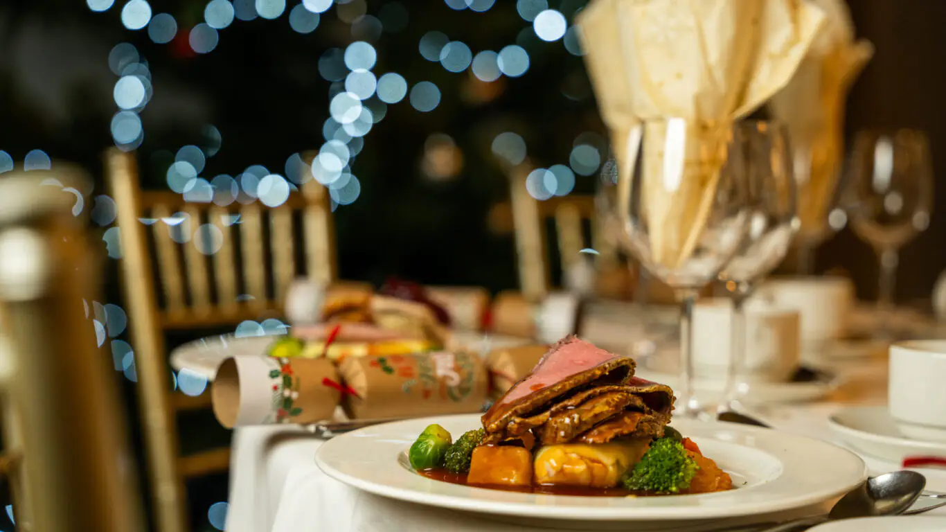A plate of roast meat with vegetables is set on a white tablecloth, alongside wine glasses, napkins, and festive decorations, with blurred lights in the background.