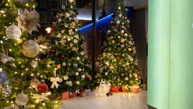 Three decorated Christmas trees with ornaments and lights, surrounded by wrapped presents, are displayed indoors on a tiled floor near a large green-lit column.