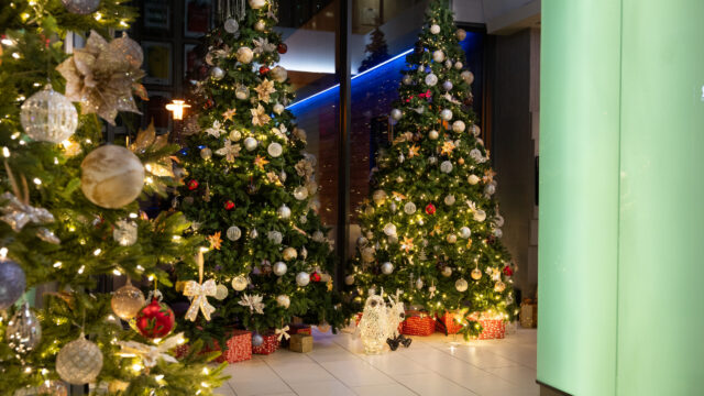 Three decorated Christmas trees with ornaments and lights, surrounded by wrapped presents, are displayed indoors on a tiled floor near a large green-lit column.