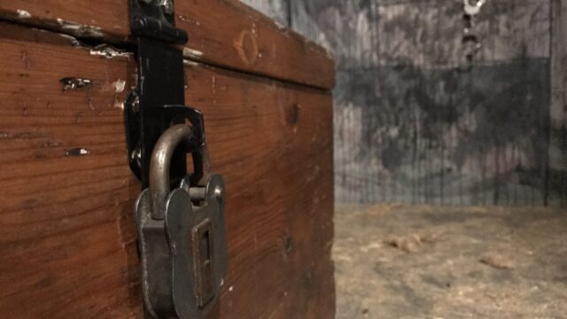 A close-up of a wooden chest secured with a large padlock, set in a dimly lit room with rough wooden floors and a blurred, textured wall in the background.