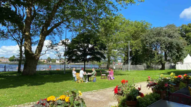 Four people sit at colorful tables on a grassy lawn near a river, surrounded by trees and flowers, on a sunny day.