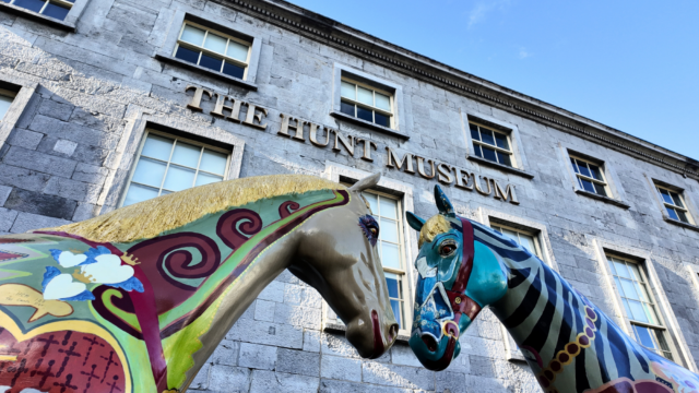Two colorful, painted horse sculptures stand outside The Hunt Museum, a stone building with large windows and signage, under a clear blue sky.
