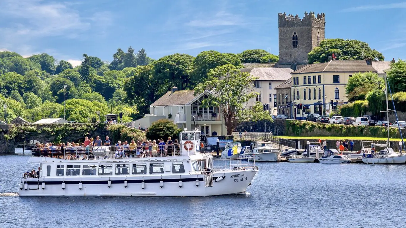 A white passenger boat sails on a river with people on deck, passing a marina, leafy trees, and historic stone buildings under a blue sky.