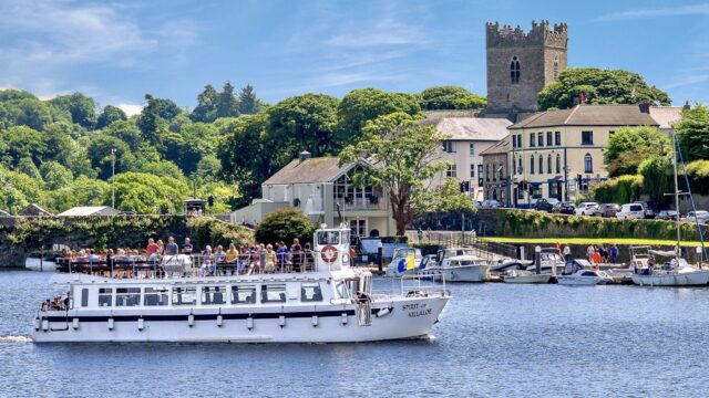 A white passenger boat sails on a river with people on deck, passing a marina, leafy trees, and historic stone buildings under a blue sky.