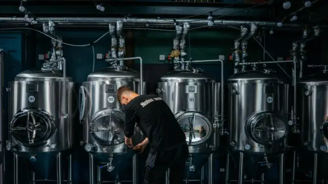 A person inspects or adjusts a valve on one of several large stainless steel brewing tanks in an industrial brewery setting.