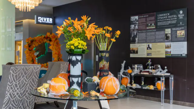 Hotel lobby decorated for autumn with pumpkins, gourds, orange flowers, and artificial cobwebs on a glass table; informational display and refreshment table in the background.