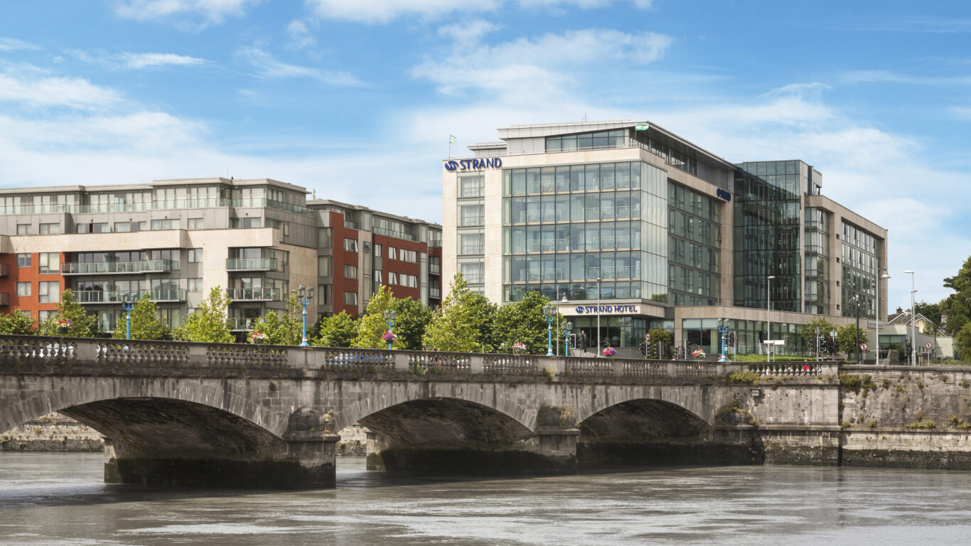 Modern glass-fronted hotel and apartment buildings stand behind a stone bridge crossing a wide river under a partly cloudy sky.