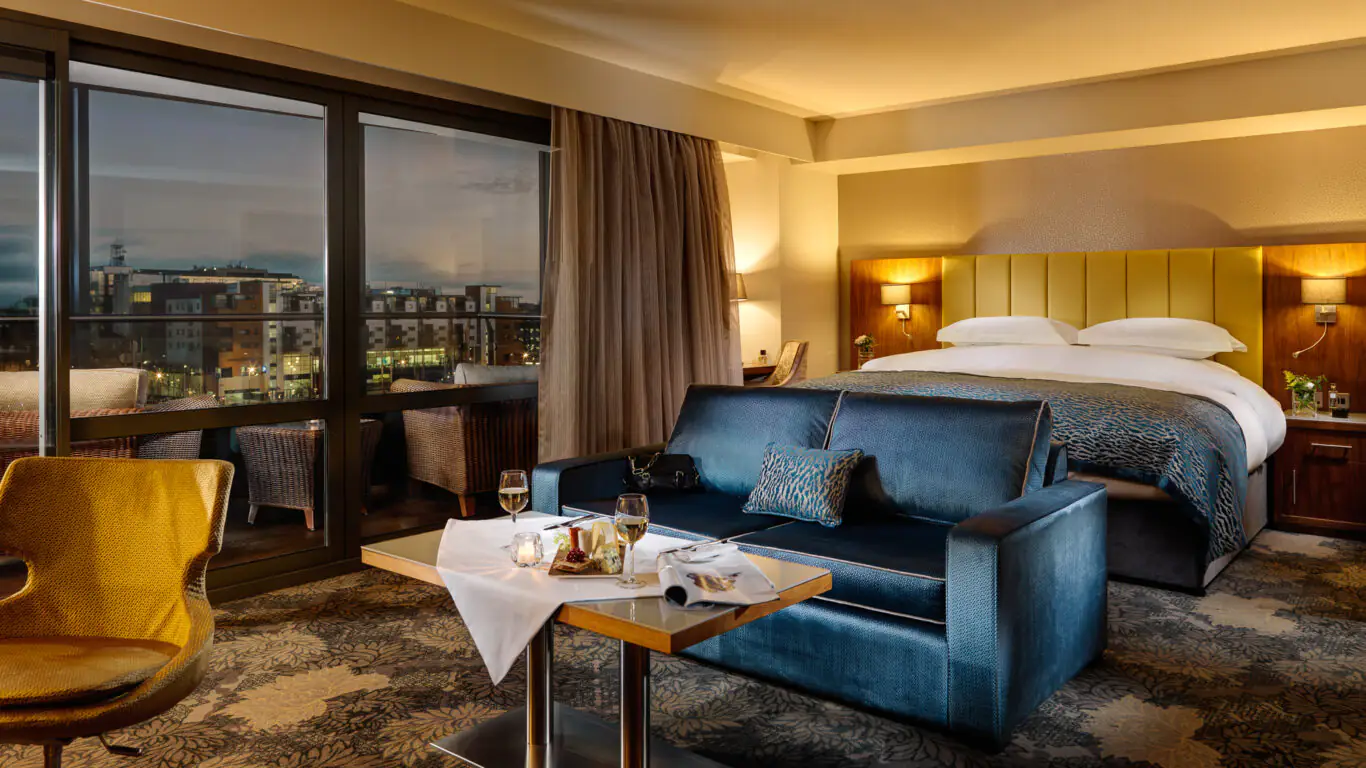 A modern hotel room with a double bed, a sofa, a table set for two with wine and food, large windows, and a city view at dusk.