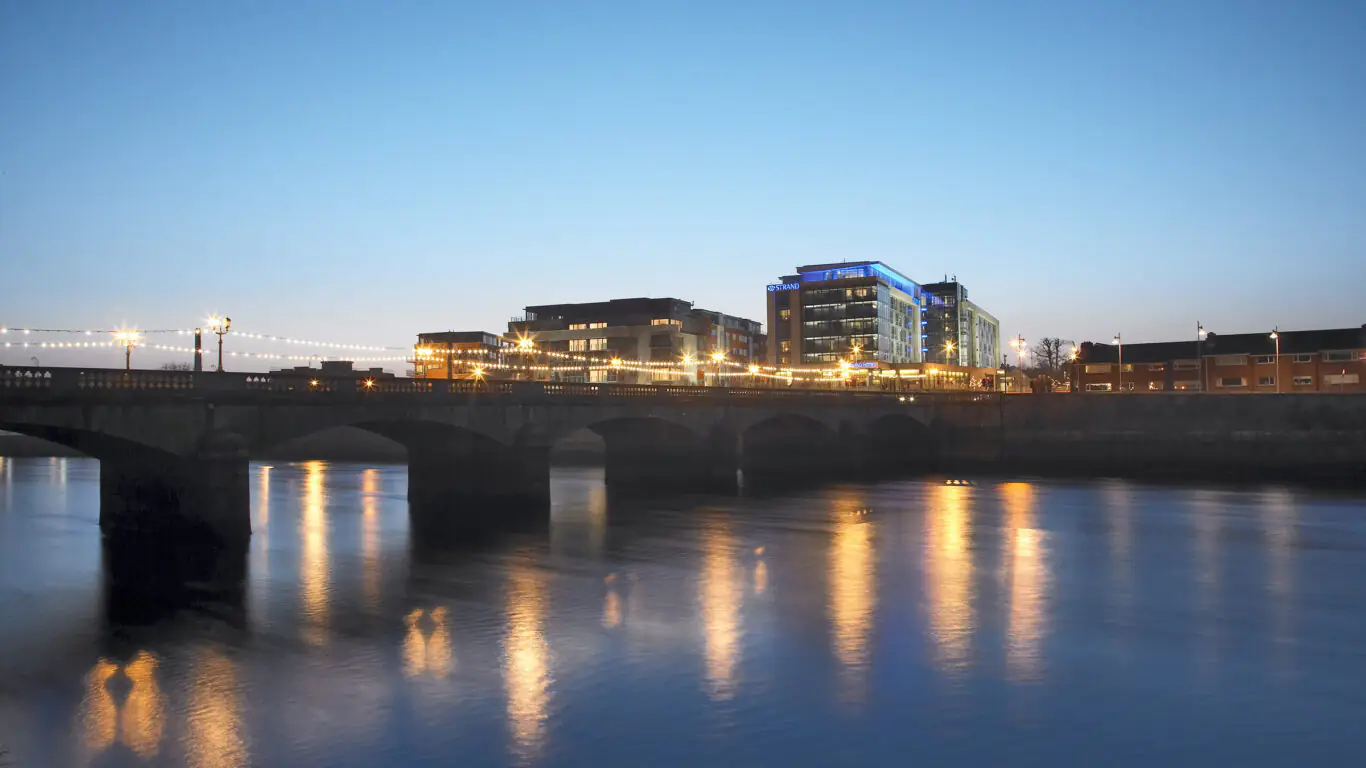 A stone bridge crosses a calm river at dusk, with modern buildings lit up in the background and lights reflecting on the water.