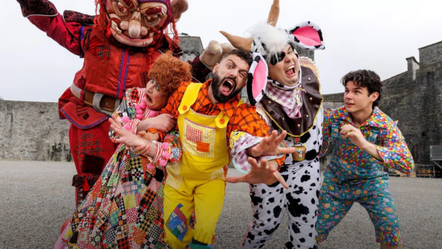 Five people in colorful, whimsical costumes pose energetically outdoors on gravel—perfect inspiration for fun Autumn Hotel Breaks, with stone walls and overcast skies in the background.