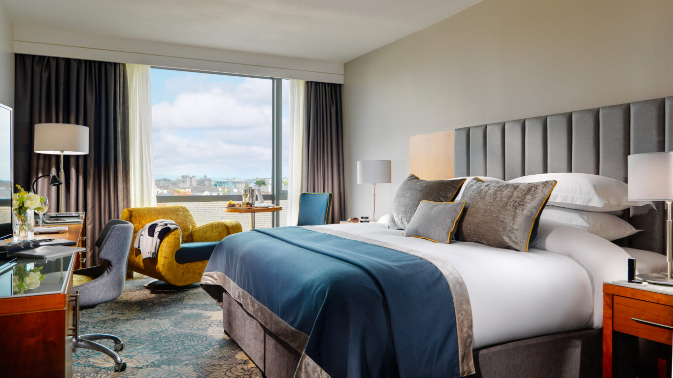 Modern hotel room with a large bed, desk, yellow armchair, and floor-to-ceiling window showing a city view under a partly cloudy sky.